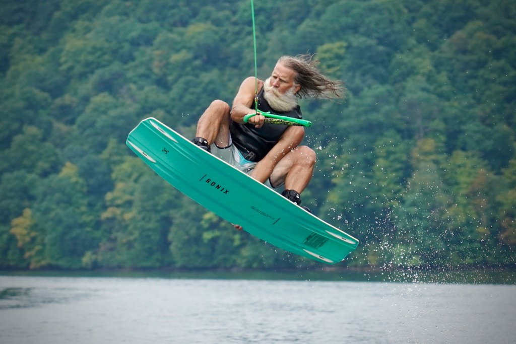 An older man with long gray hair and a beard is wakeboarding on a lake, holding onto a green fishing rod while in mid-air.