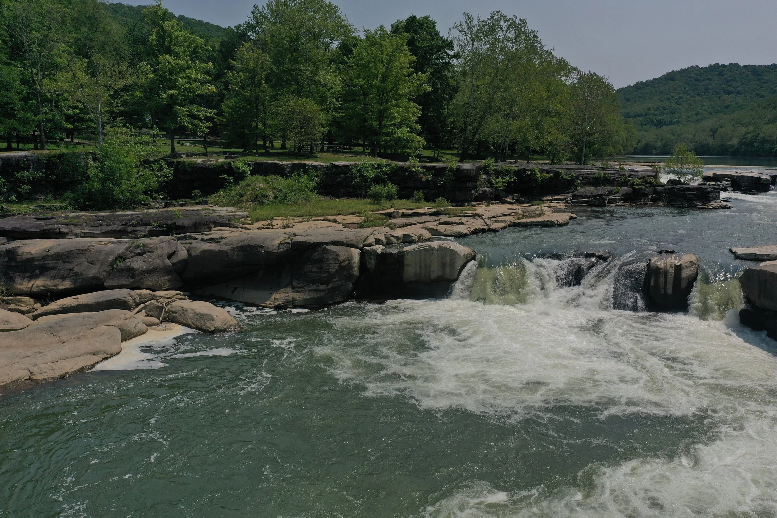 A flowing river with rocks and small waterfalls, surrounded by green trees and hills in the background.