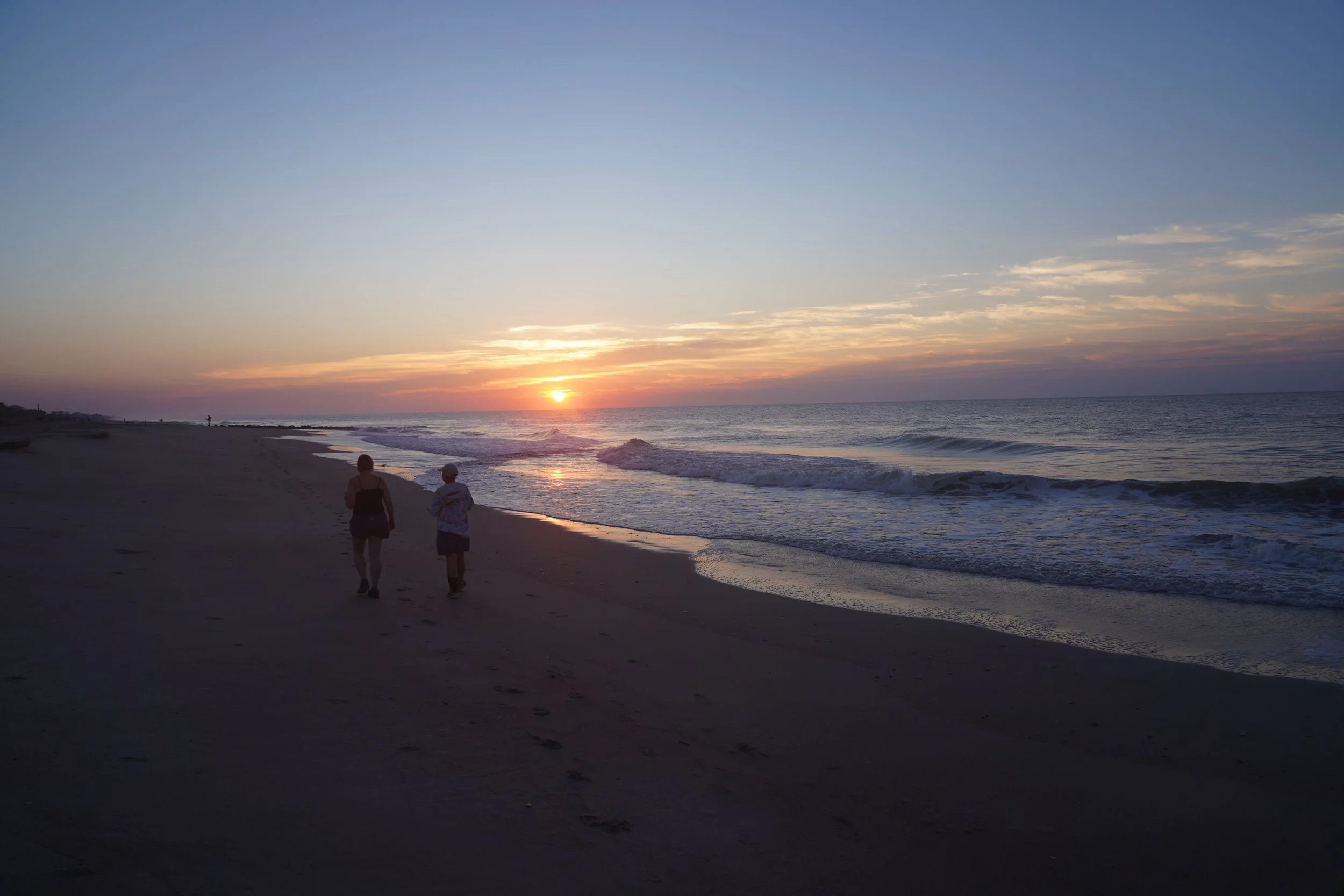 Two people walking along a beach at sunset, with the ocean waves gently crashing ashore and a colorful sky.