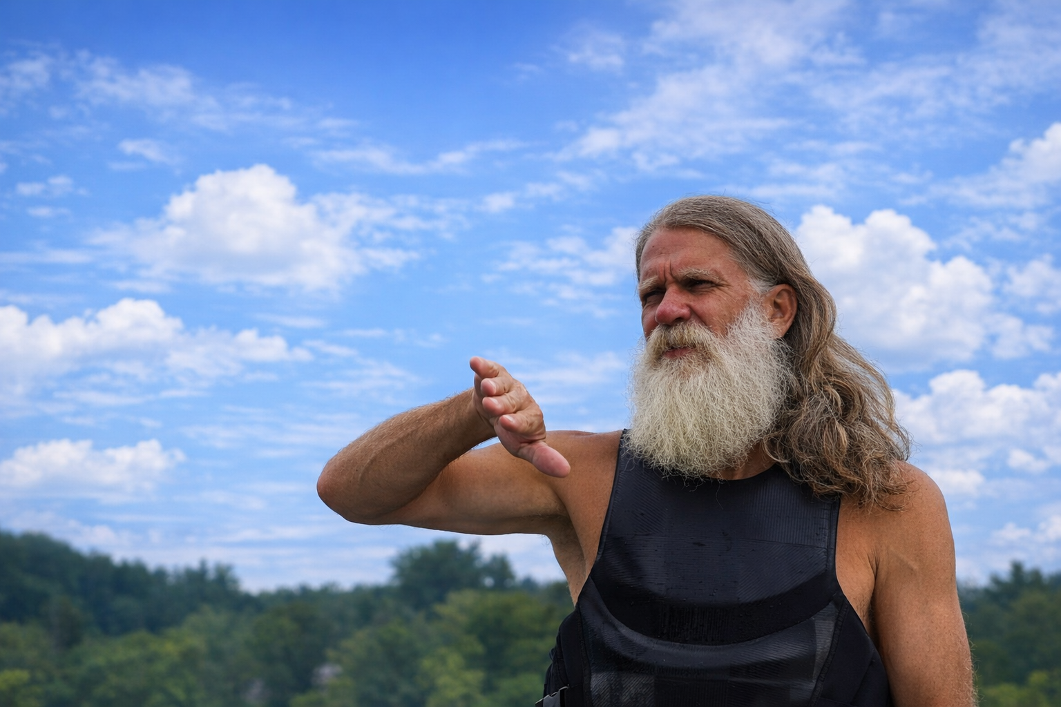 An elderly man with a long white beard and long hair standing outdoors on a sunny day, wearing a black sleeveless shirt, making a gesture with his hand under his chin, with a background of blue sky and trees.