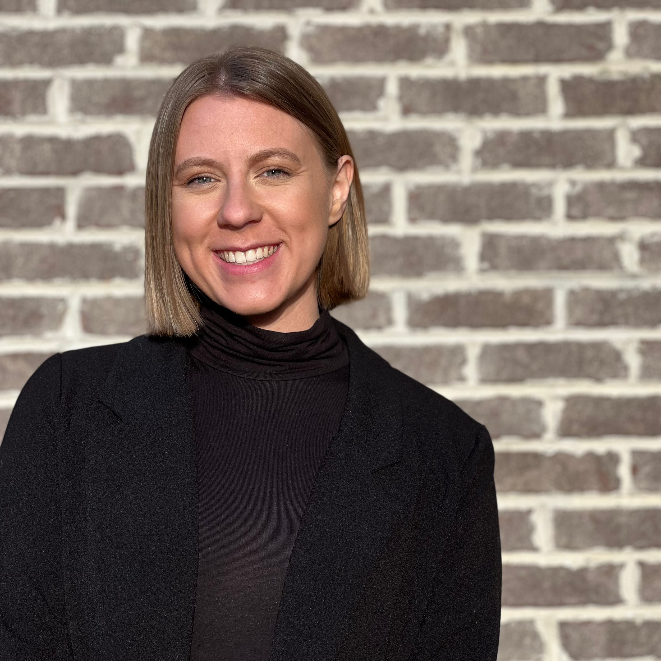 Lauren, the founder of The SEL Studio. Image is of a woman with shoulder-length light brown hair smiling, wearing a black turtleneck and blazer, standing in front of a brick wall.