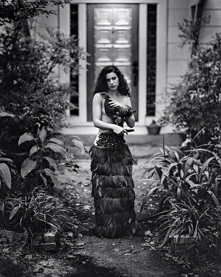 A woman with curly hair in an elaborate feathered skirt and sleeveless top holding a chicken in her hand, standing on a garden path in front of a house with a door, with plants on both sides.
