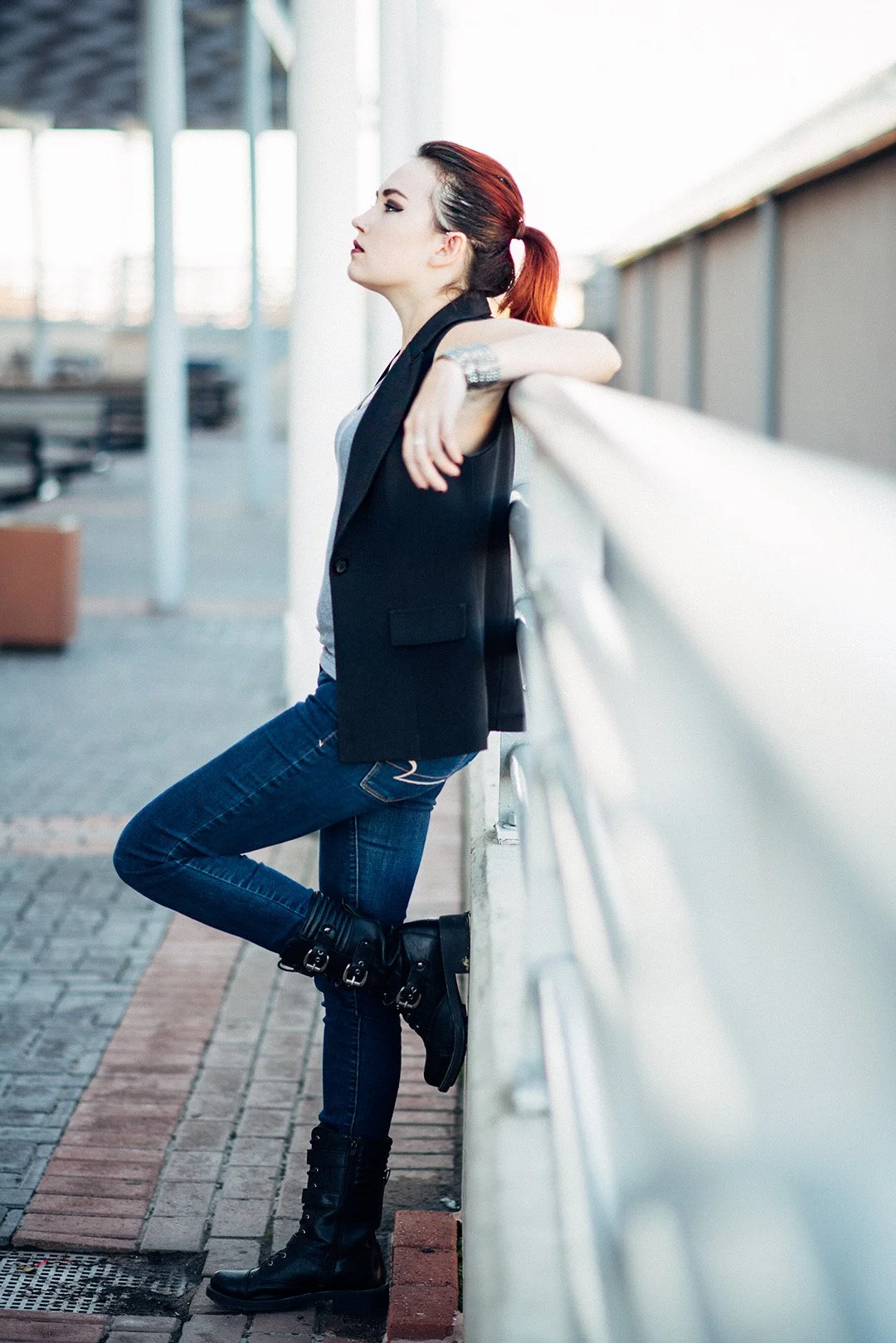 A woman with red hair styled in a ponytail leaning against a railing outdoors, wearing a black vest, gray top, blue jeans, and black boots.