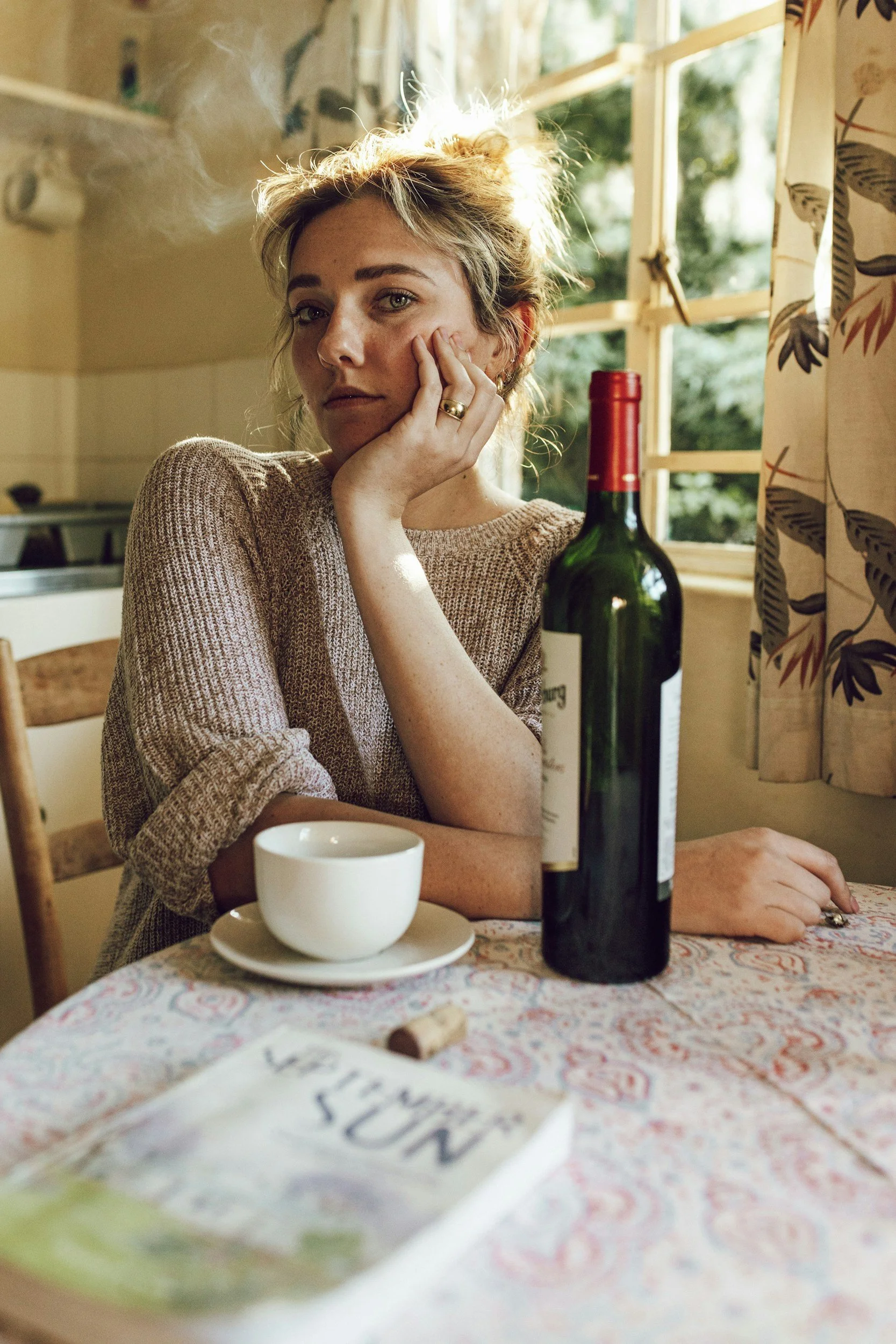 A woman with light hair and fair skin sitting at a table in a cozy kitchen, resting her face on her hand, with a thoughtful expression. There is a bottle of red wine, a white coffee cup on a saucer, a cork, and a magazine titled 'VISIT' on the table. Sunlight streams through the window behind her, illuminating the scene.