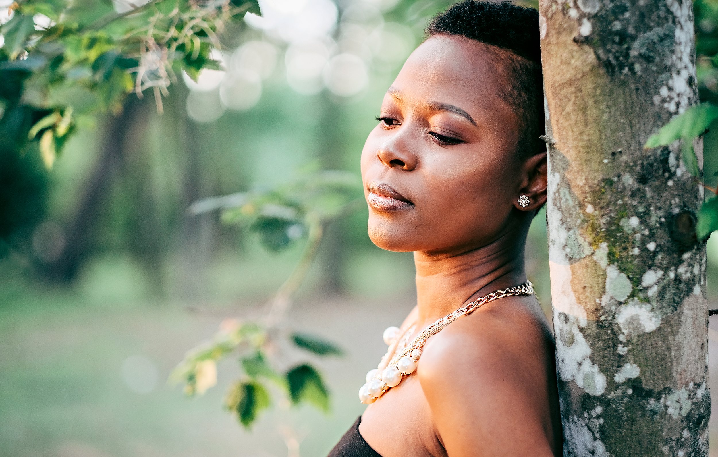 A woman with short hair and jewelry leans against a tree outdoors, looking contemplative.