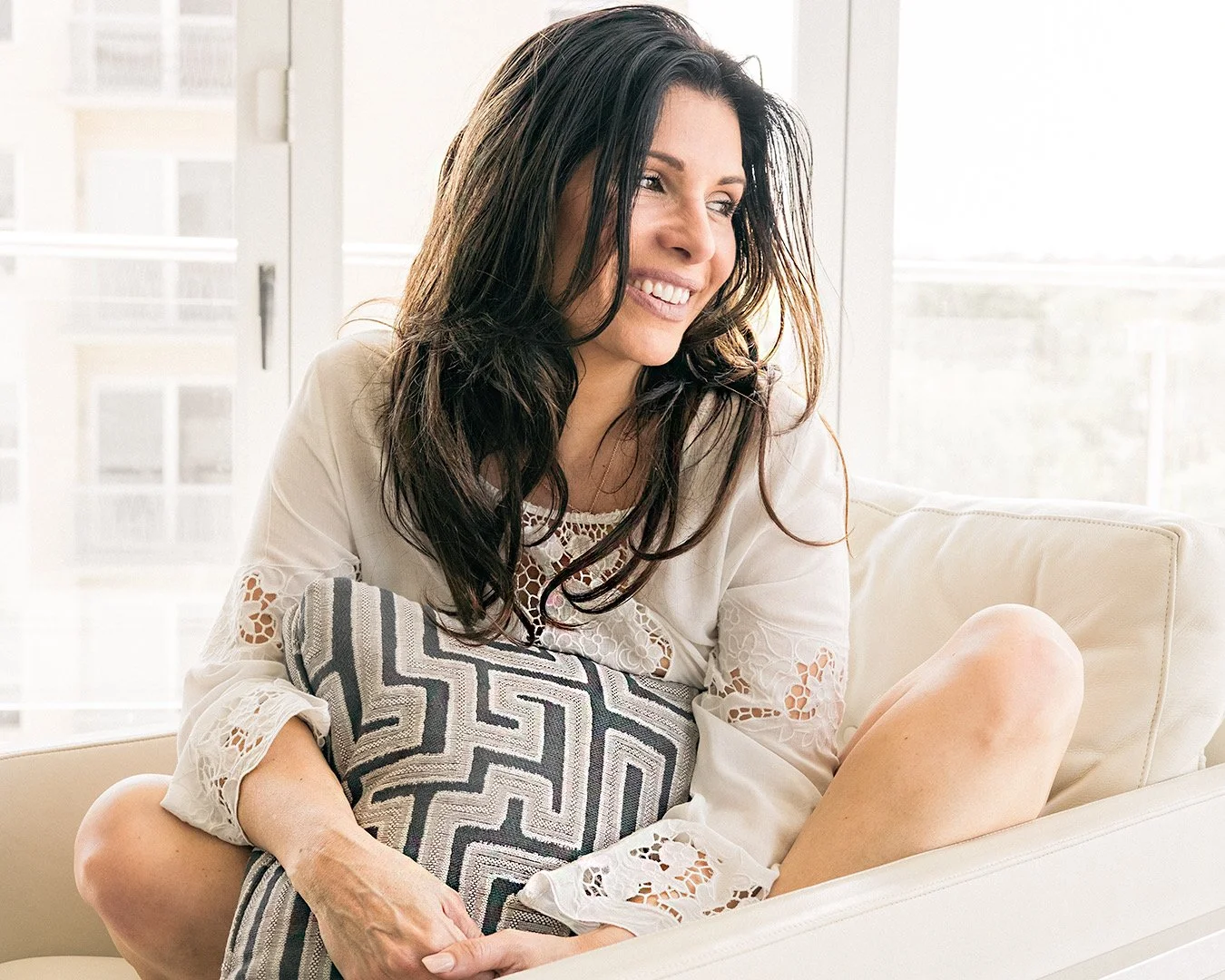 Woman smiling on a cream-colored couch holding a decorative pillow in a bright living room.