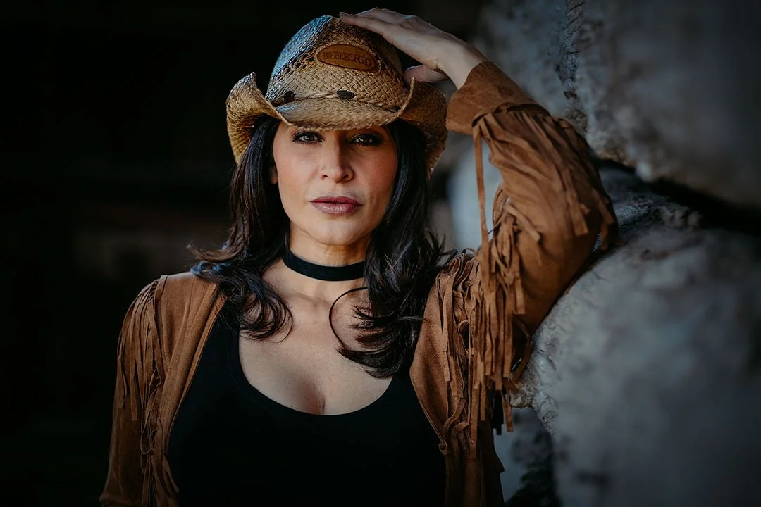 A woman wearing a cowboy hat, brown fringed jacket, and black top, standing against a rocky background.