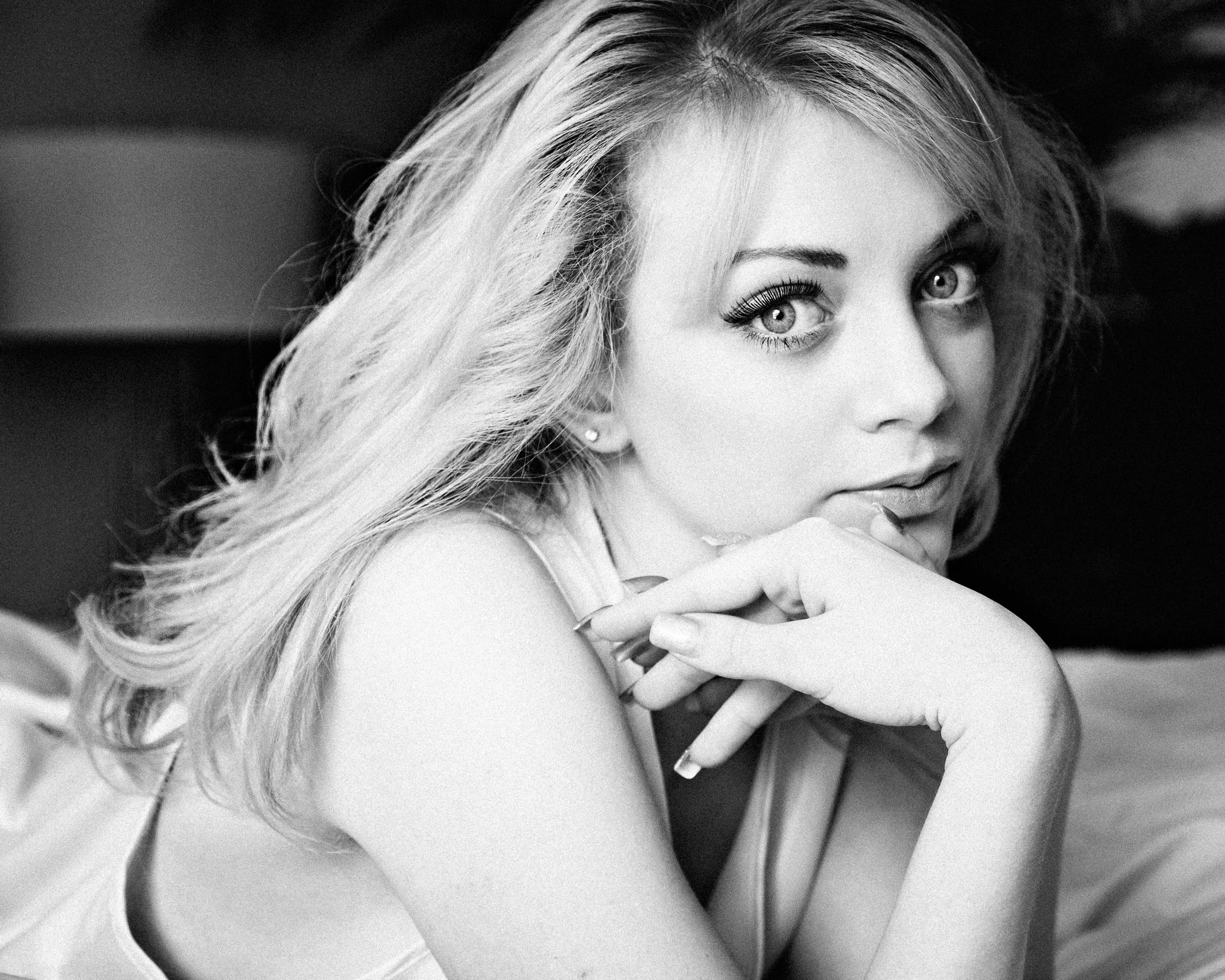 A woman with light-colored hair, wearing a sleeveless top, resting her chin on her hand and looking into the camera with a soft expression.