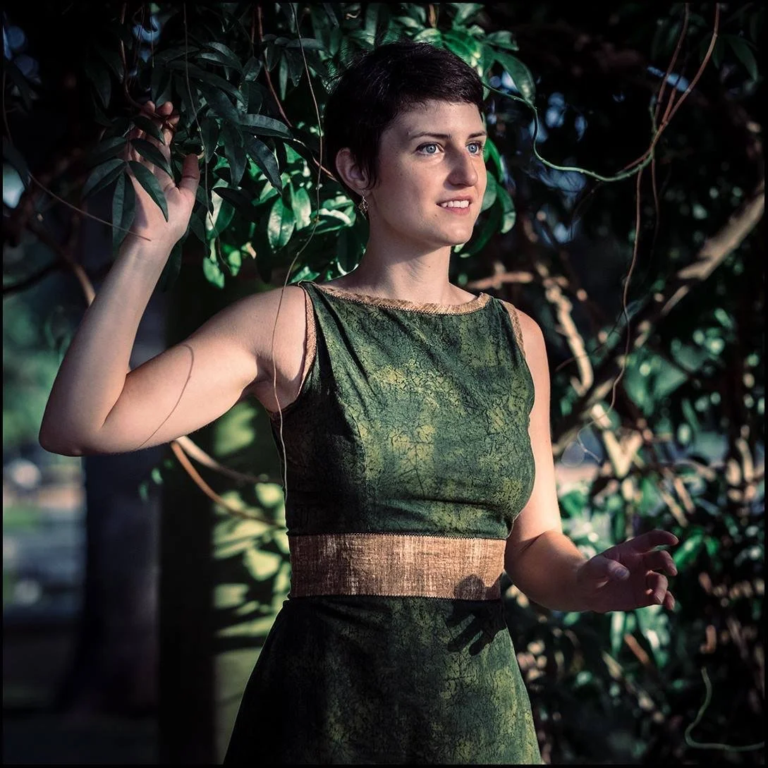 A woman with short dark hair and blue eyes standing outdoors among green foliage, wearing a sleeveless green dress with a brown waistband, looking into the distance.
