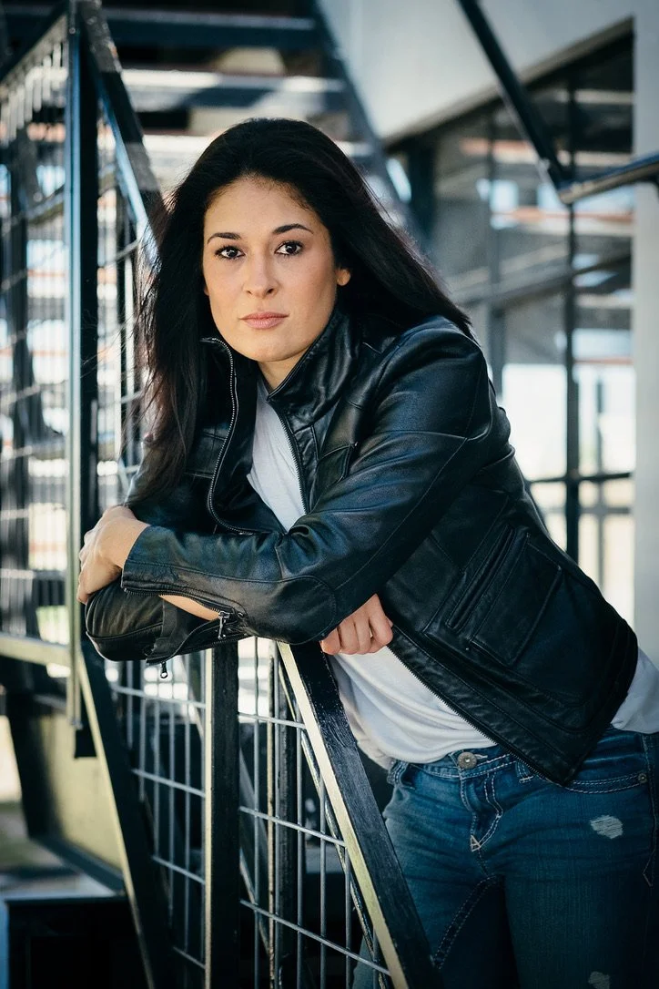 A woman with long dark hair wearing a black leather jacket and white shirt, leaning on a shopping cart at an indoor location with metal stairs or racks in the background.