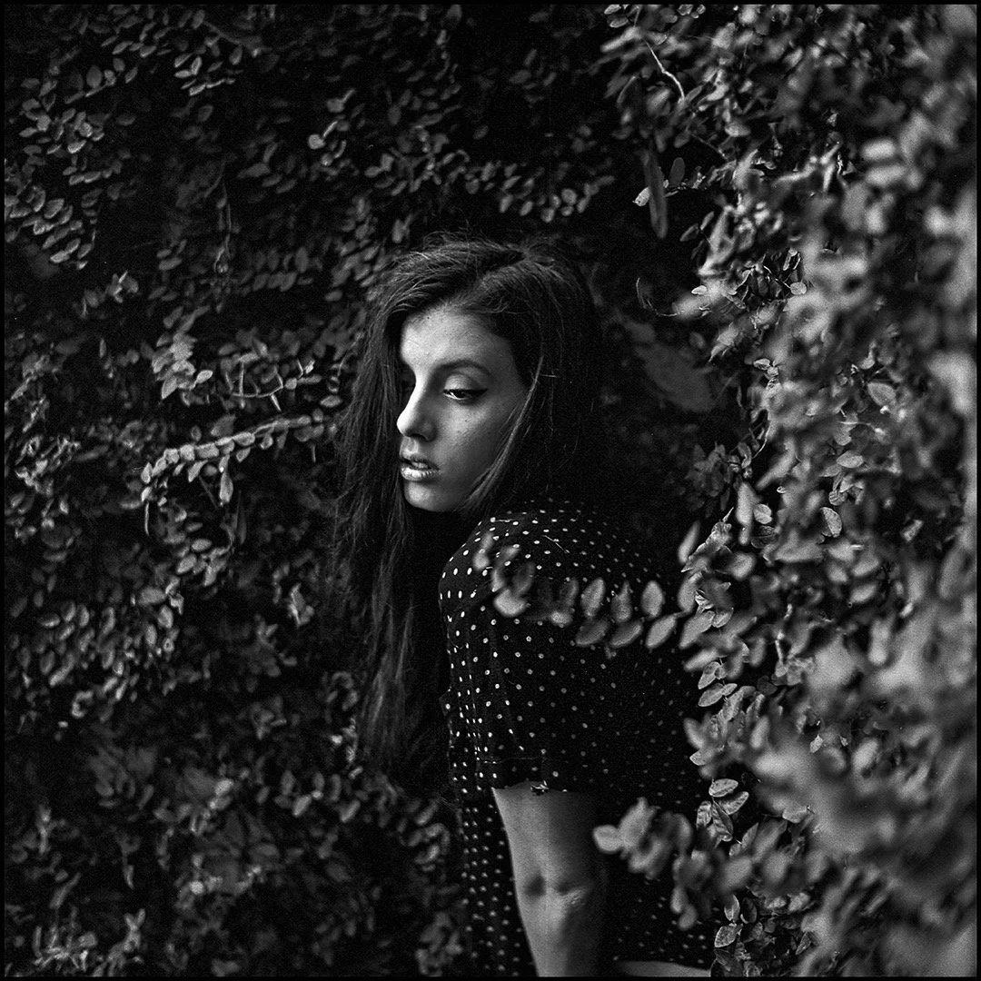 Black and white photo of a woman with long dark hair and a polka dot dress, standing among dense foliage, looking downward with a contemplative expression.