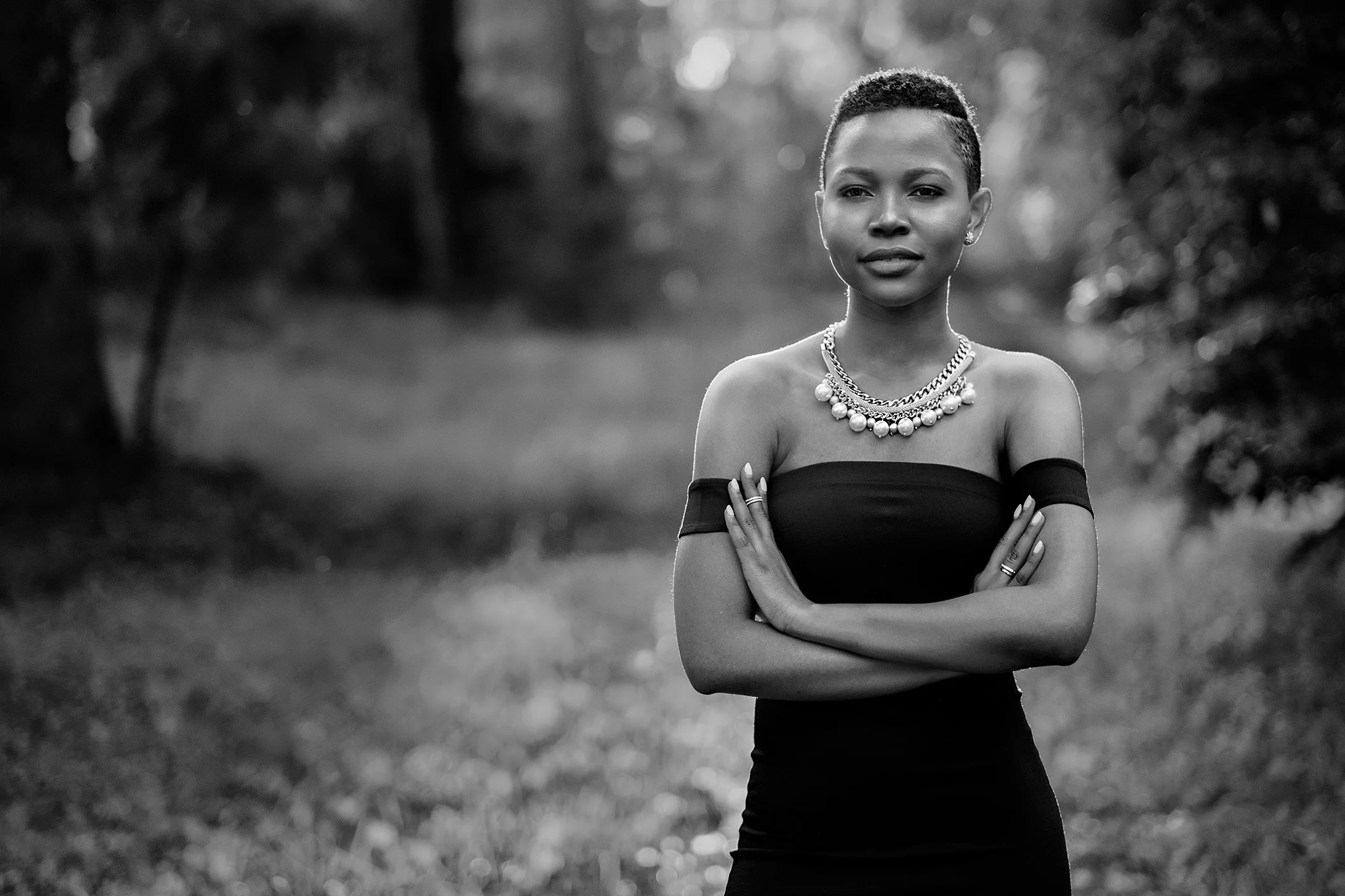 A woman standing outdoors in a natural setting during daylight, wearing a strapless black dress and layered necklaces, with her arms crossed.
