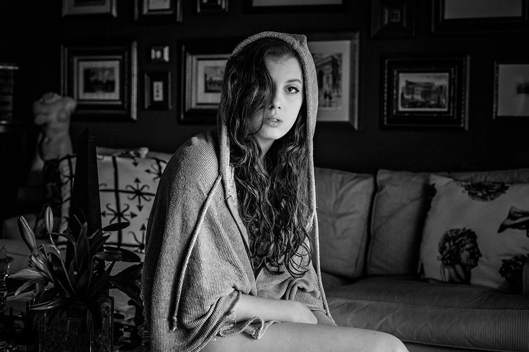 Black and white photo of a young woman with long wavy hair, wearing a hooded sweater, sitting on a couch in a room decorated with framed pictures on the wall and pillows on the couch.