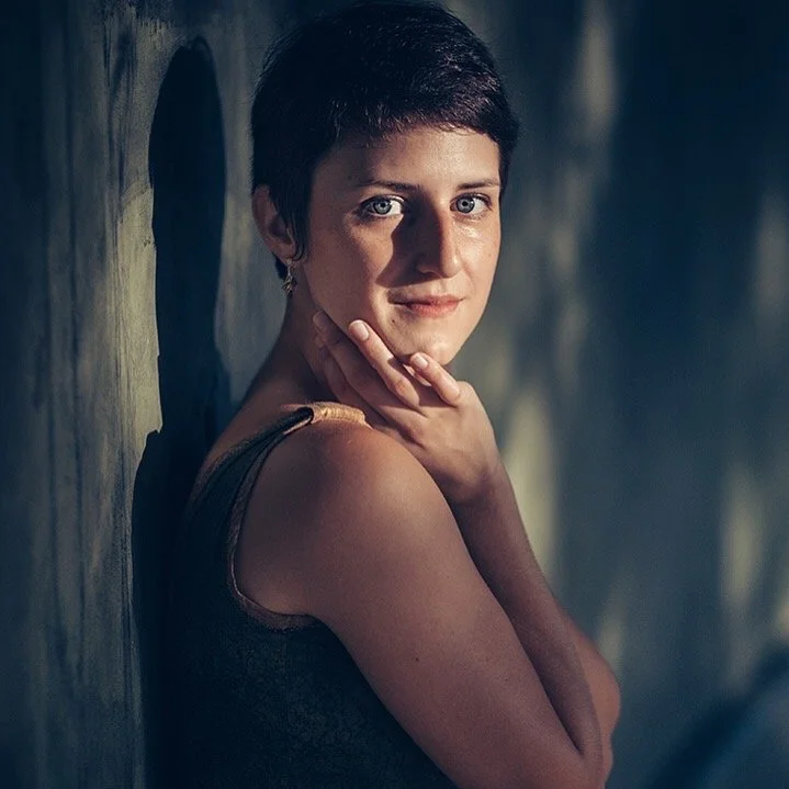 A woman with short dark hair and fair skin, wearing a sleeveless top, standing against a wall with shadows cast on it.