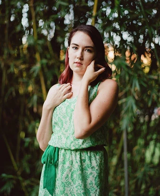 Young woman with reddish hair in a green lace dress standing outdoors among tall green plants with blurred background of foliage and sunlight.