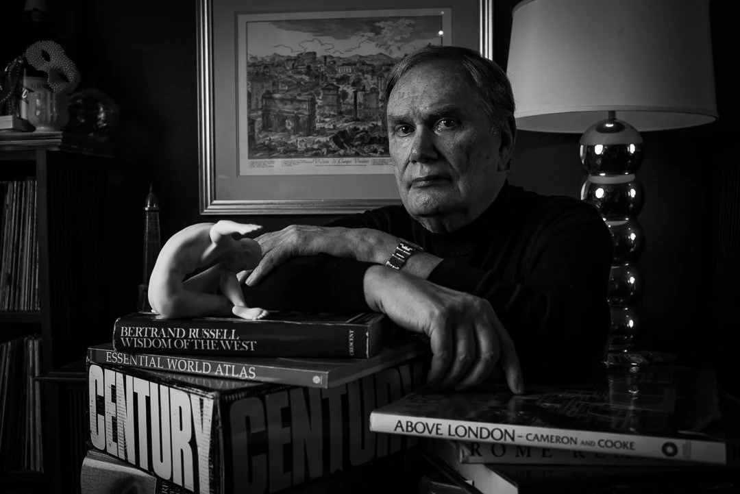 A black and white photo of an elderly man sitting at a table with books and a sculpture. There is a framed picture on the wall behind him and a lamp on his right.