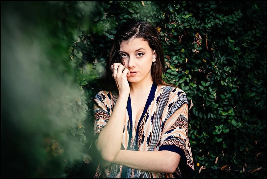 A young woman with dark hair, wearing a patterned top, standing in front of a dense green leafy background, with one hand near her face and looking at the camera.