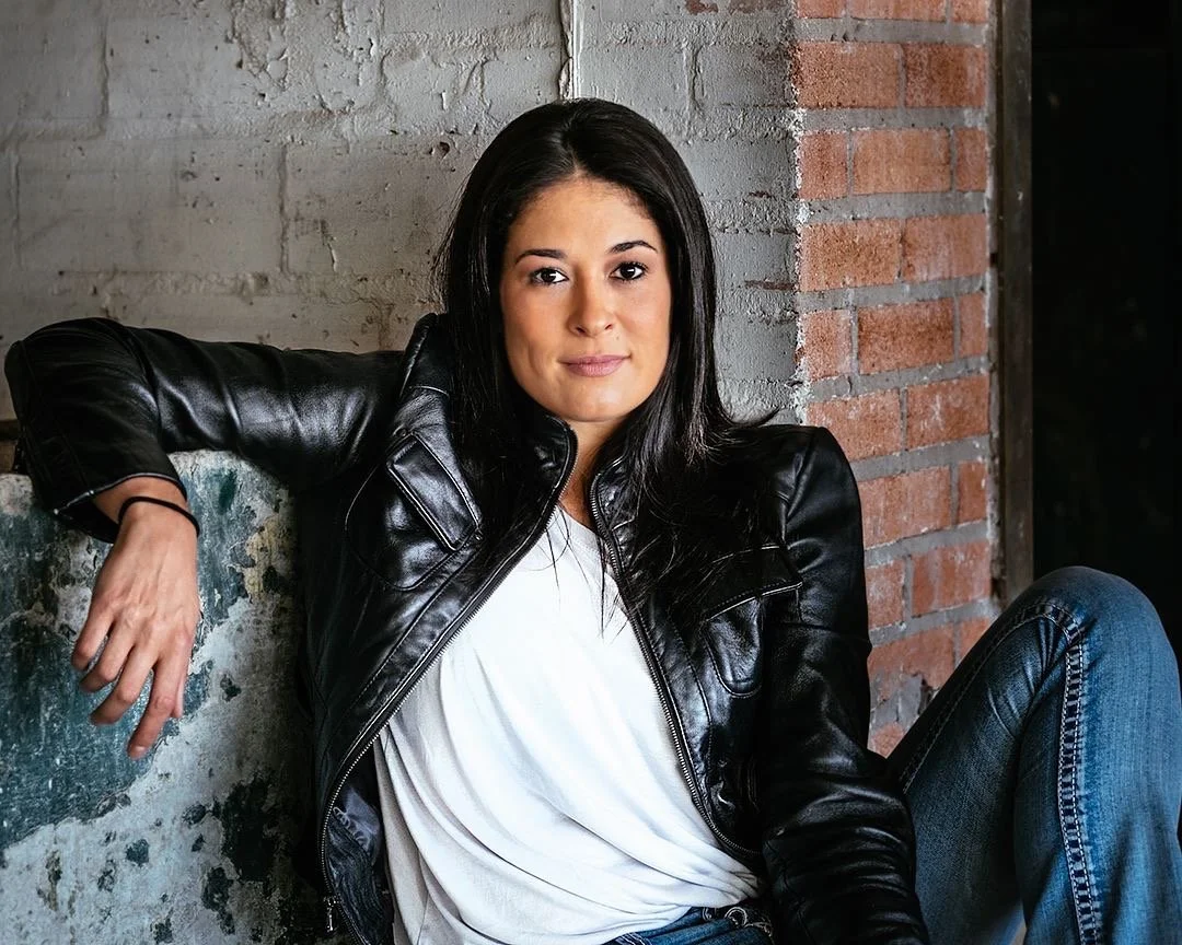 A woman with long dark hair wearing a black leather jacket and blue jeans sitting on a concrete ledge in front of a brick and concrete wall.