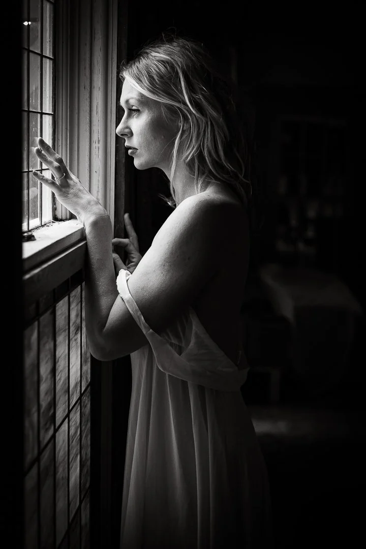 A black and white photo of a woman with wavy hair looking out a window, touching the window with one hand, wearing a light, loose dress with off-shoulder sleeves.