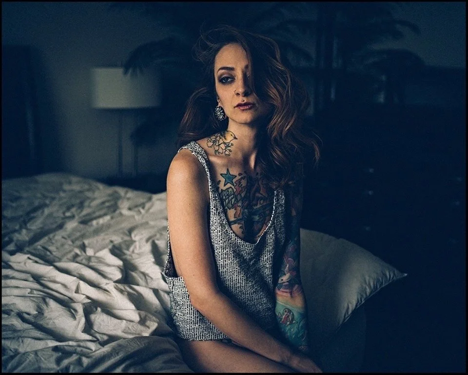 A woman with long red hair and visible tattoos sitting on a bed in a dimly lit room, wearing a sleeveless top.