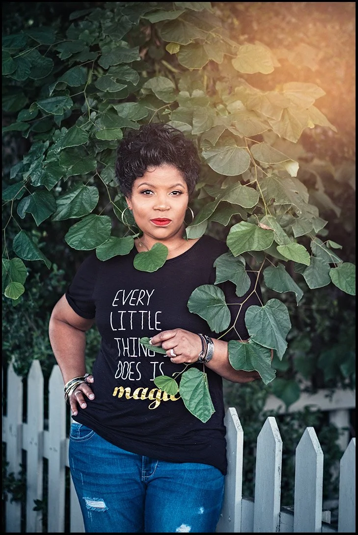 A woman with dark hair and red lipstick stands outdoors among green leaves and a white picket fence, wearing a black T-shirt with the words 'Every little thing does is magique' and denim jeans.