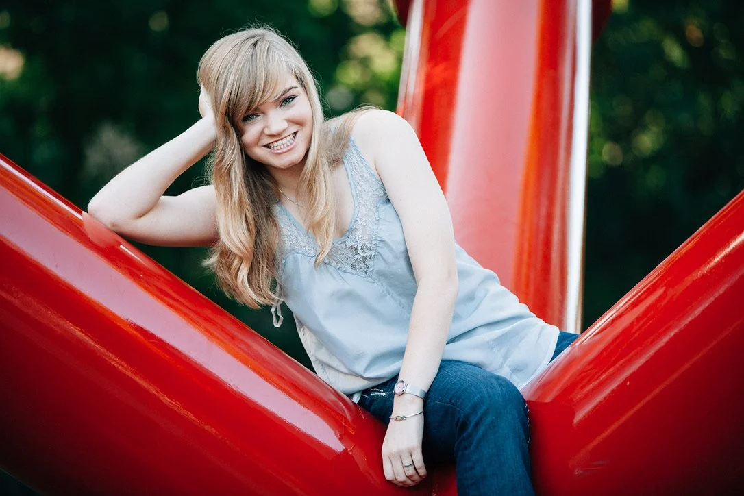 A young woman with long, wavy blonde hair smiling and sitting on a red playground structure outdoors, with a blurred green background.