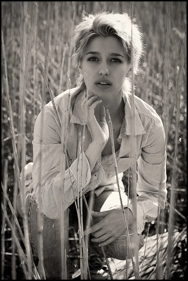 A woman with short blonde hair crouching among tall grass in a field, looking directly at the camera in black and white.