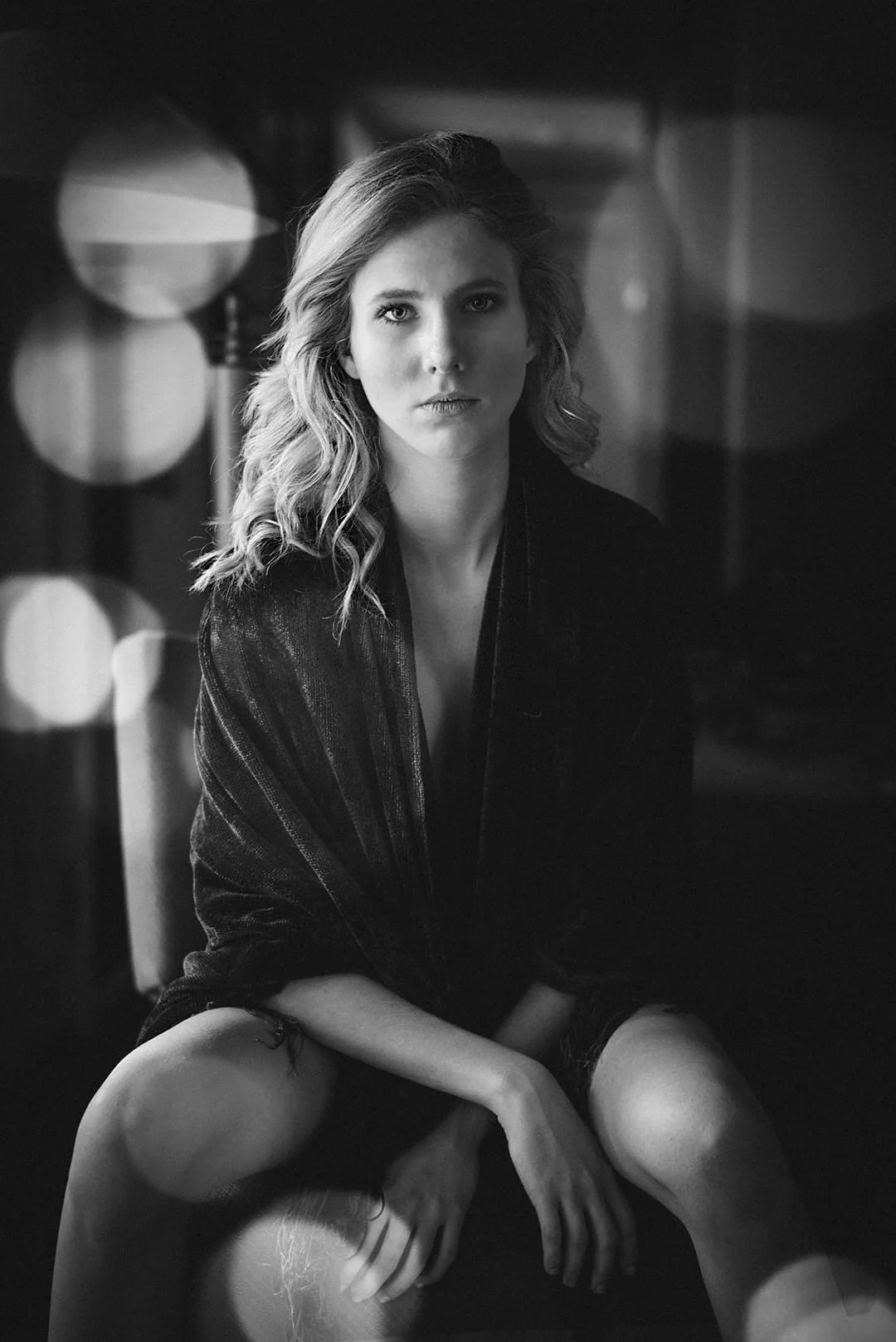 A black and white portrait of a young woman sitting with her legs apart, wearing a dark robe or dress, with loose wavy hair, looking directly into the camera.