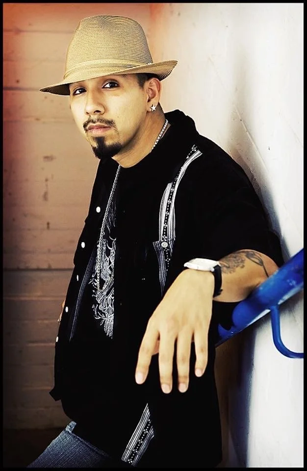 A man wearing a beige fedora hat, black jacket with silver zippers, and a black graphic T-shirt, leaning against a white wall with a blue railing.