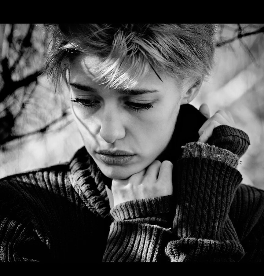 A black and white portrait of a young person with short, tousled hair, looking downward with their hand near their face, wearing a striped, cozy sweater.