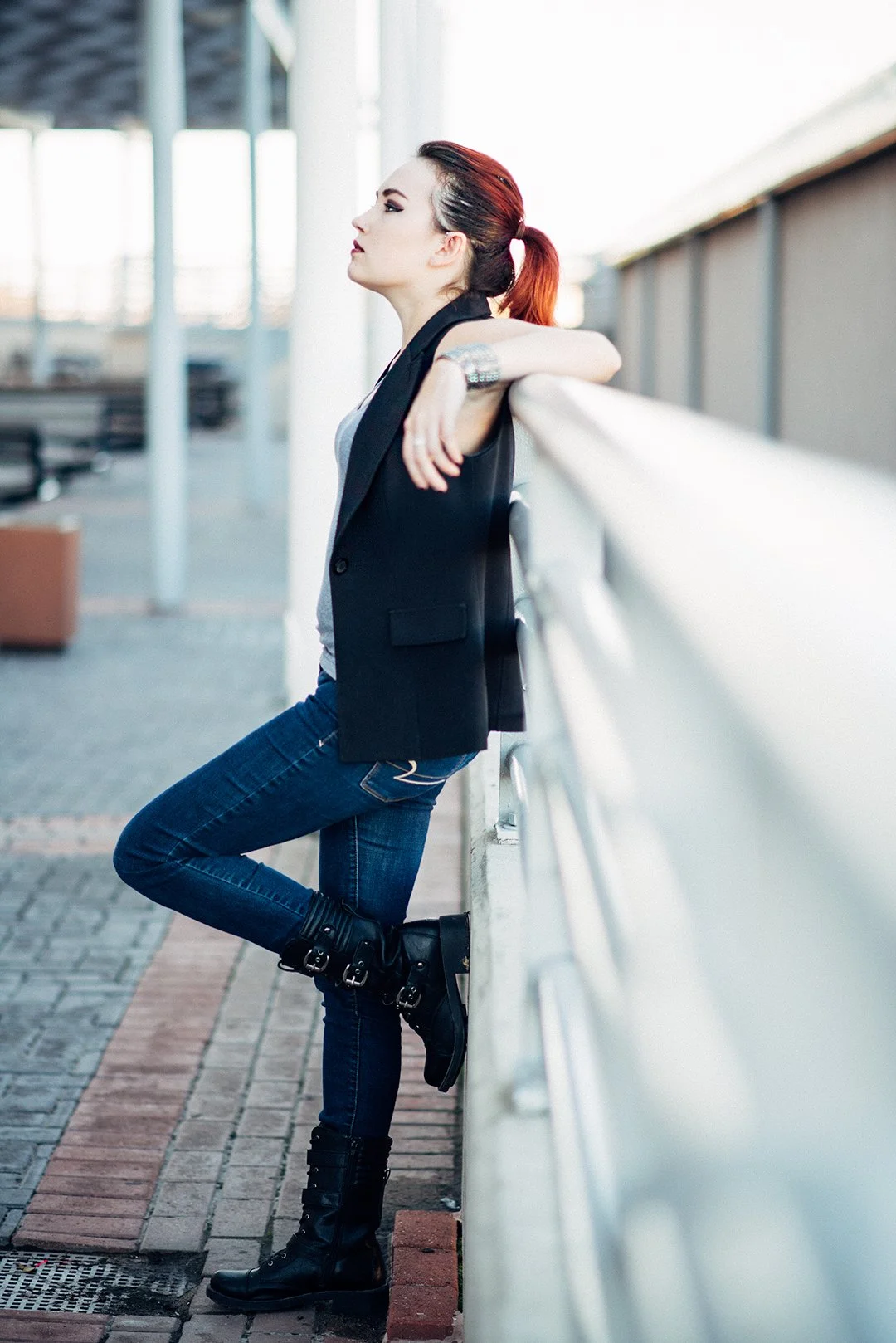 A woman with red hair tied back, wearing a black vest, gray top, blue jeans, and black boots, leaning against a white railing on an outdoor walkway.