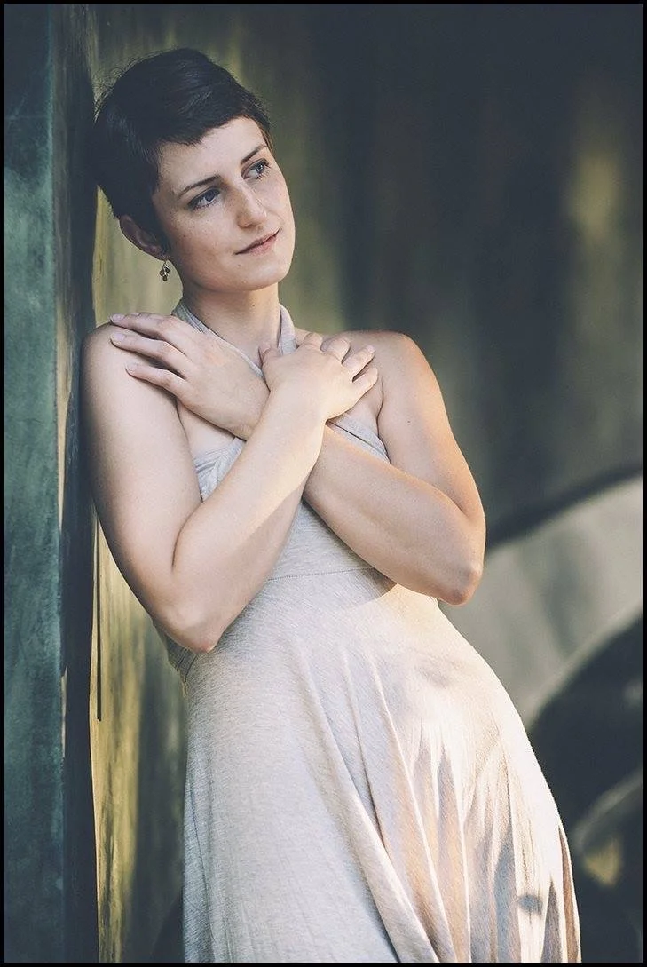 A woman with short dark hair and fair skin, wearing a light-colored dress, stands with her back against a wall, arms crossed over her chest, gazing thoughtfully into the distance.