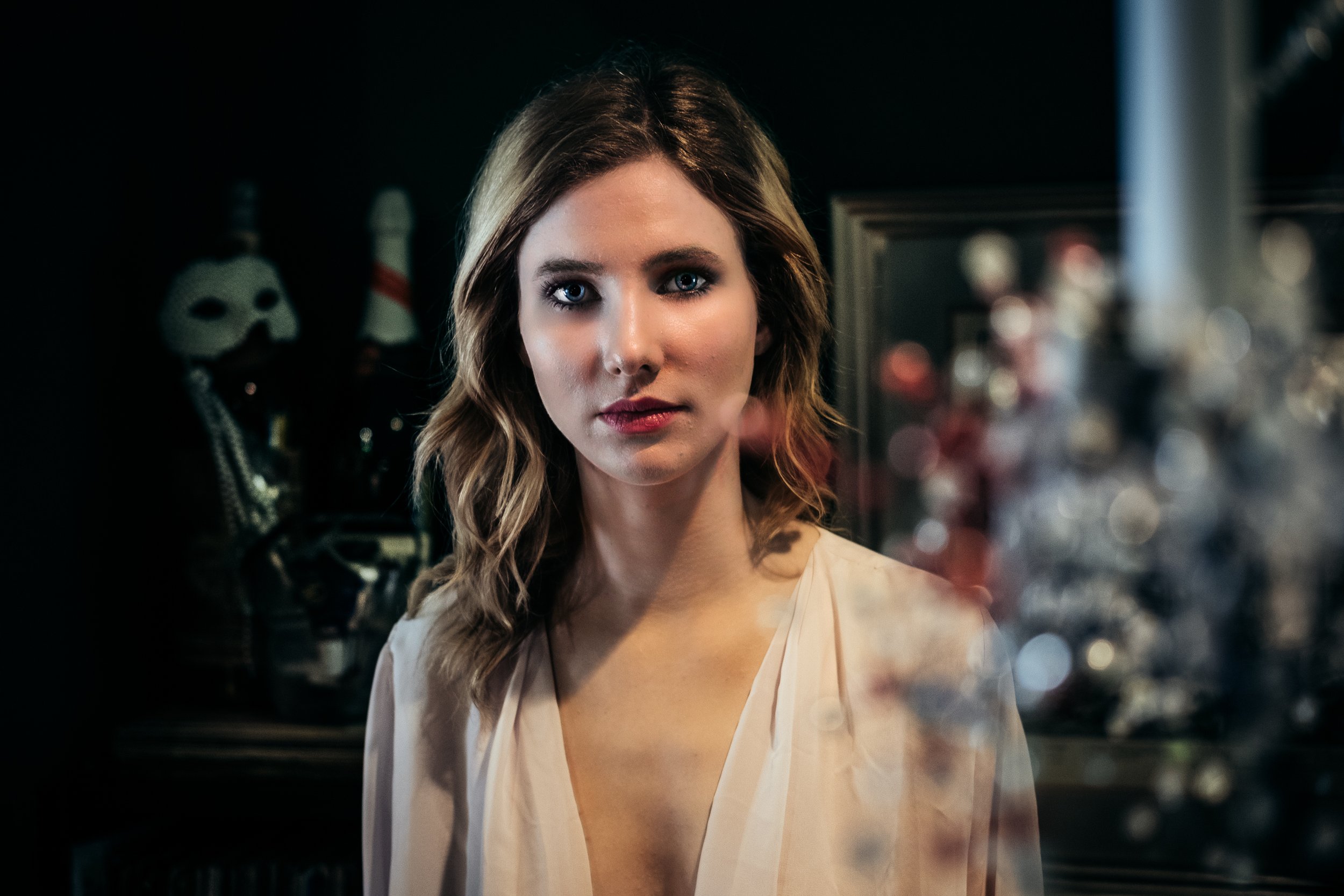 A woman with wavy brown hair and blue eyes looking directly at the camera, wearing a light-colored blouse, in a dimly lit room with masks and framed mirrors in the background.