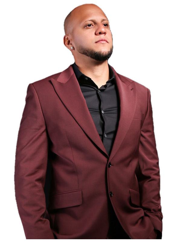 A man with a shaved head and a goatee wearing a maroon suit and black shirt stands against a dark background.