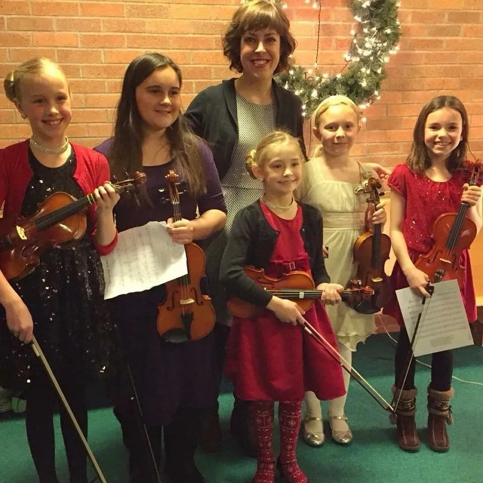 A group of six girls holding violins and a music sheet, standing with a woman in front of a brick wall decorated with a Christmas wreath with lights, suggesting a holiday concert setting.