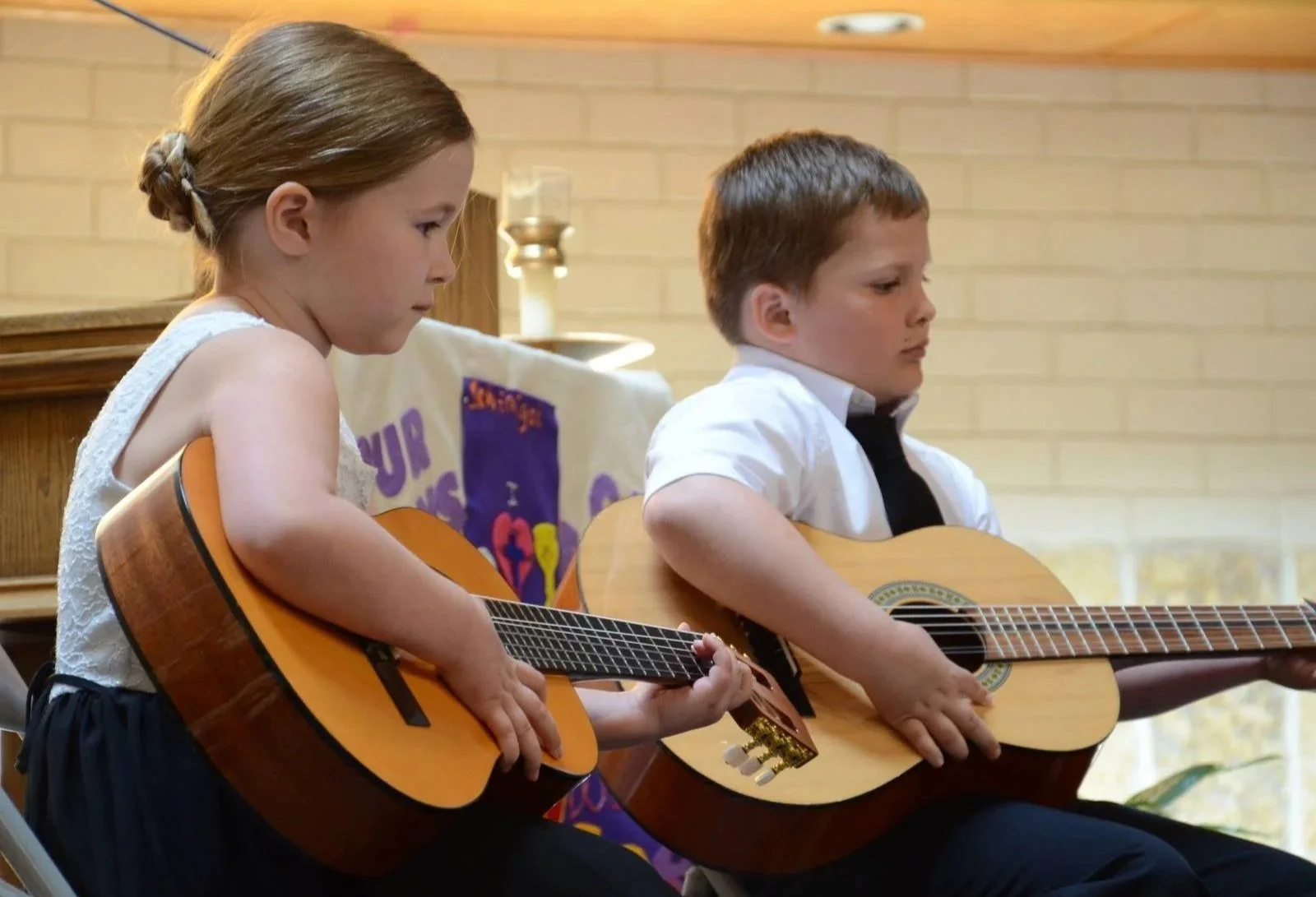 A young girl and boy sitting side by side, playing acoustic guitars during a performance or practice in an indoor setting.