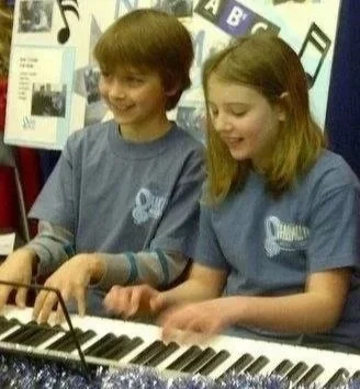 Two children, a boy and a girl, playing a keyboard together in a classroom.