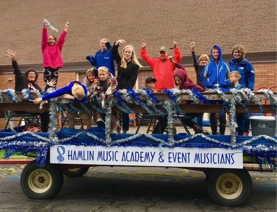 Children and adults on a decorated float at a parade, with a sign reading 'Hamlin Music Academy & Event Musicians,' celebrating a festive event.