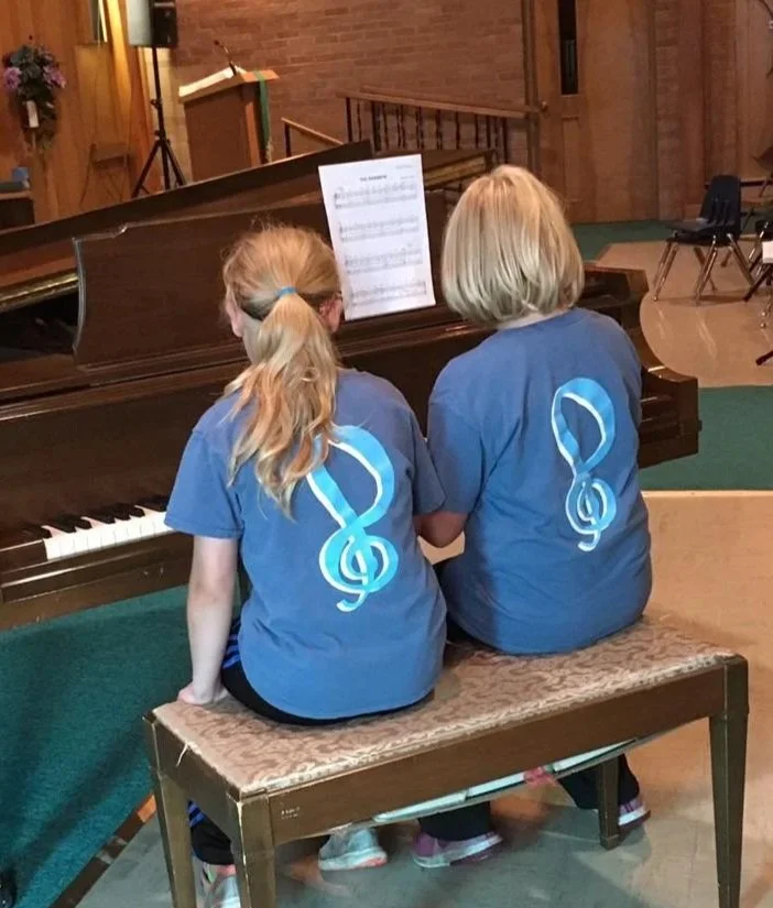 Two young girls sitting side by side at a piano, both wearing matching blue T-shirts with a treble clef and musical note design on the back, in a room with wood-paneled walls and music stands.