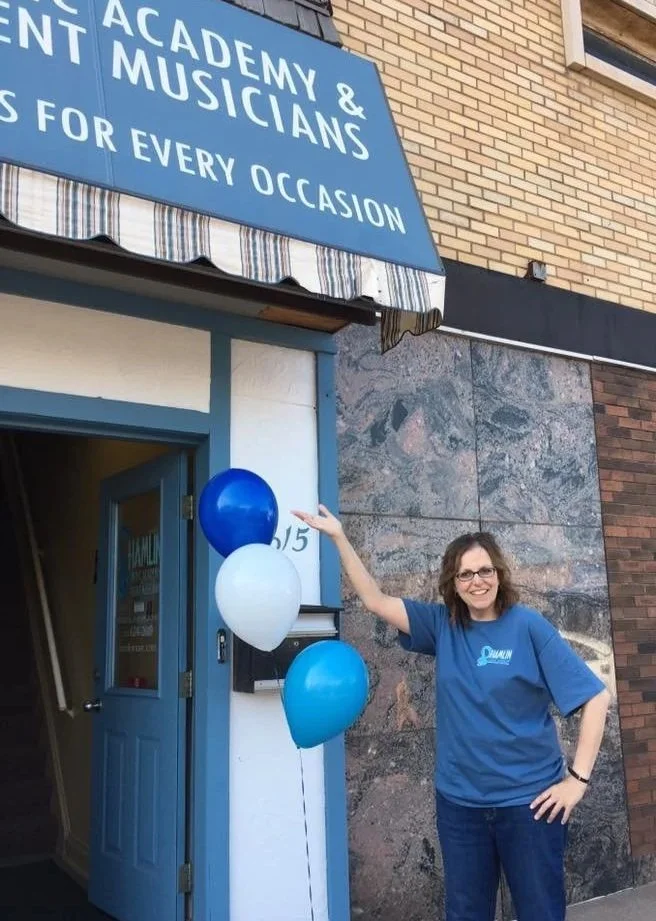 A woman with glasses and a blue shirt standing outside a building decorated with blue, white, and light blue balloons, pointing at the building's address with her right hand. The building has a sign that reads 'Academy & Talent Musicians for every oc