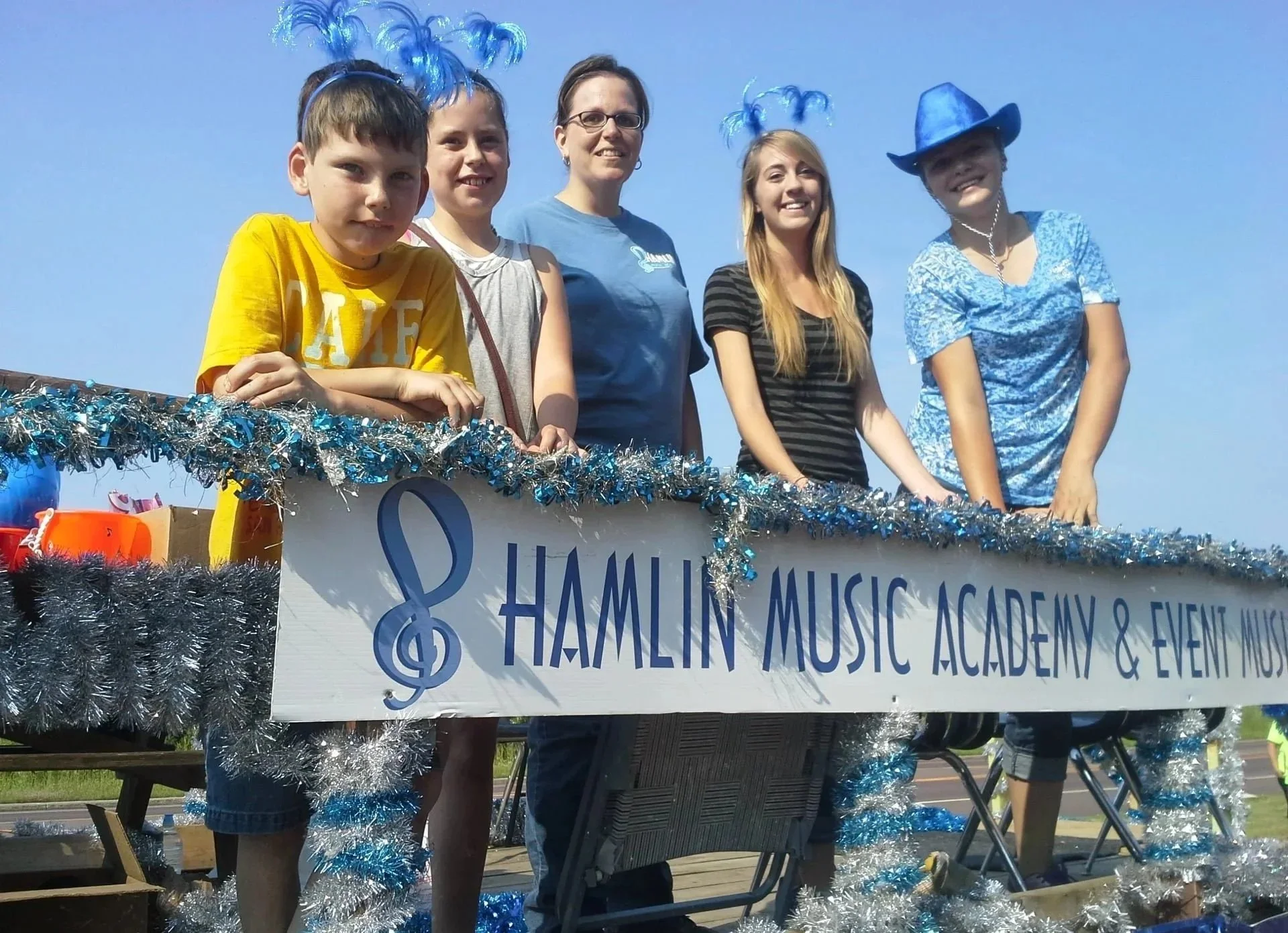 A group of people standing on a decorated float with a sign reading "Hamlin Music Academy & Event Music" during a parade, with blue and silver tinsel and streamers.