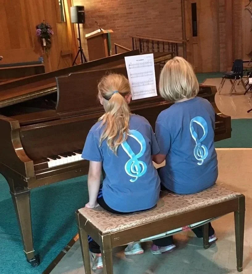 Two children, a girl with blonde hair in a ponytail and a girl with short blonde hair, sitting on a bench in front of a piano, both wearing matching blue shirts with a large treble clef symbol on the back, inside a room that appears to be a church or