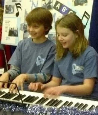 A boy and a girl playing a keyboard together in a classroom or music room, smiling and wearing matching blue shirts.