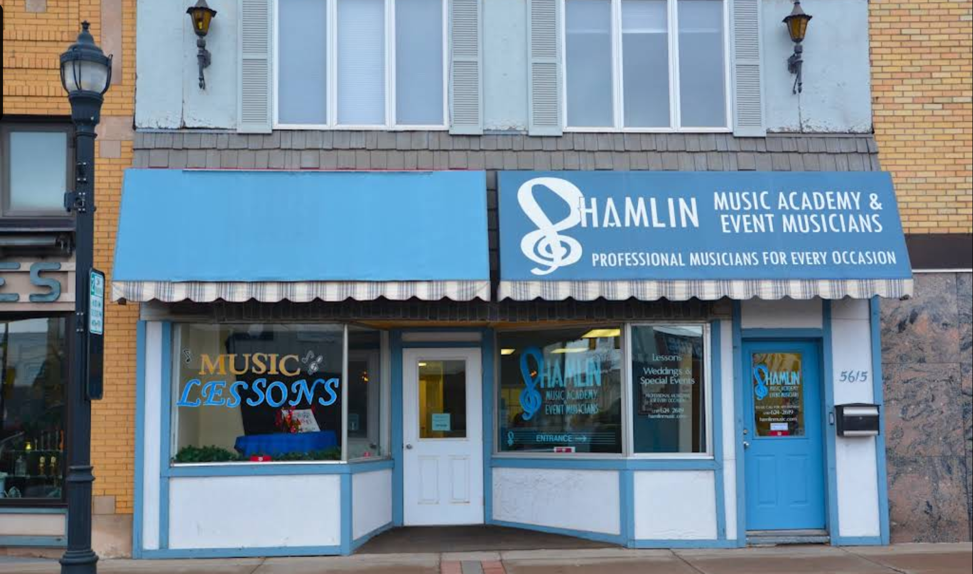 Exterior of a music school with a blue awning, sign reading 'Hamlin Music Academy & Event Musicians,' and windows displaying 'Music Lessons' in the storefront.