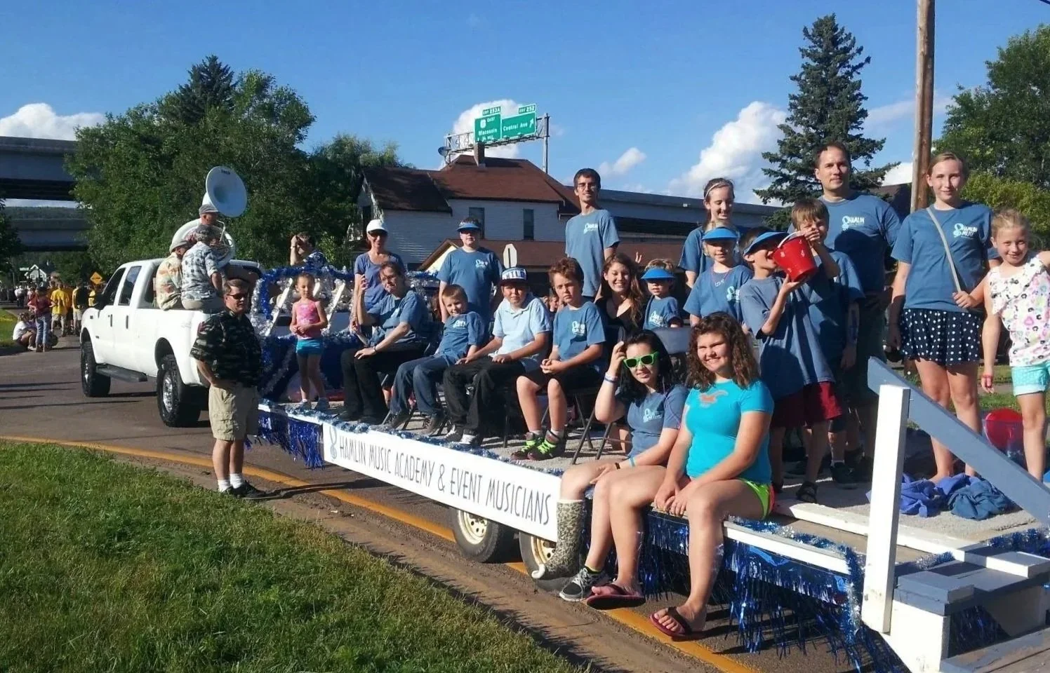 Group of children and adults on a parade float with a sign that reads 'Hamlin Music Academy & Event Musicians', wearing matching blue shirts, some with sunglasses, posing for a photo on a sunny day.