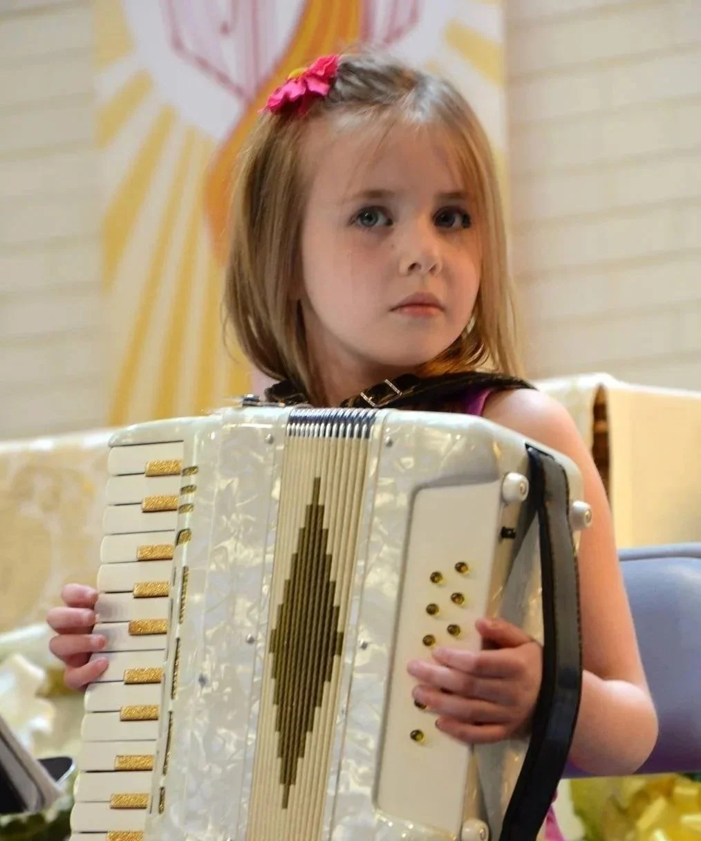 A young girl with shoulder-length blonde hair, wearing a pink hair bow, holds an accordion in a room with light-colored walls and a decorative background.