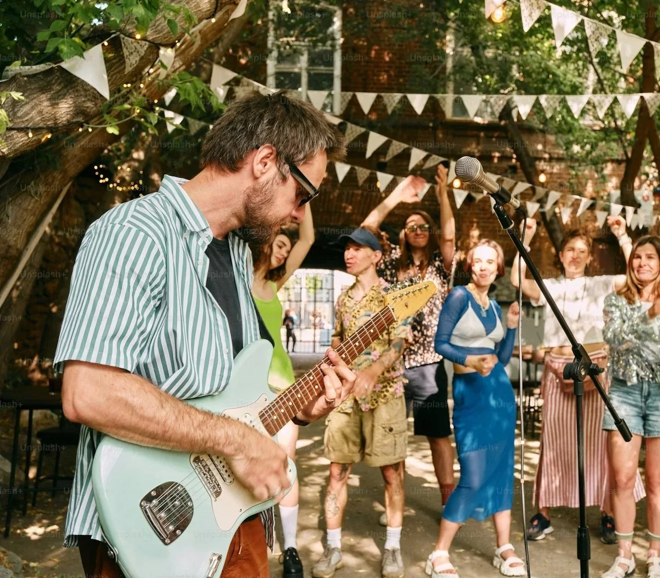 A man playing an electric guitar at an outdoor celebration, with a group of people dancing and cheering in the background under string lights and bunting.