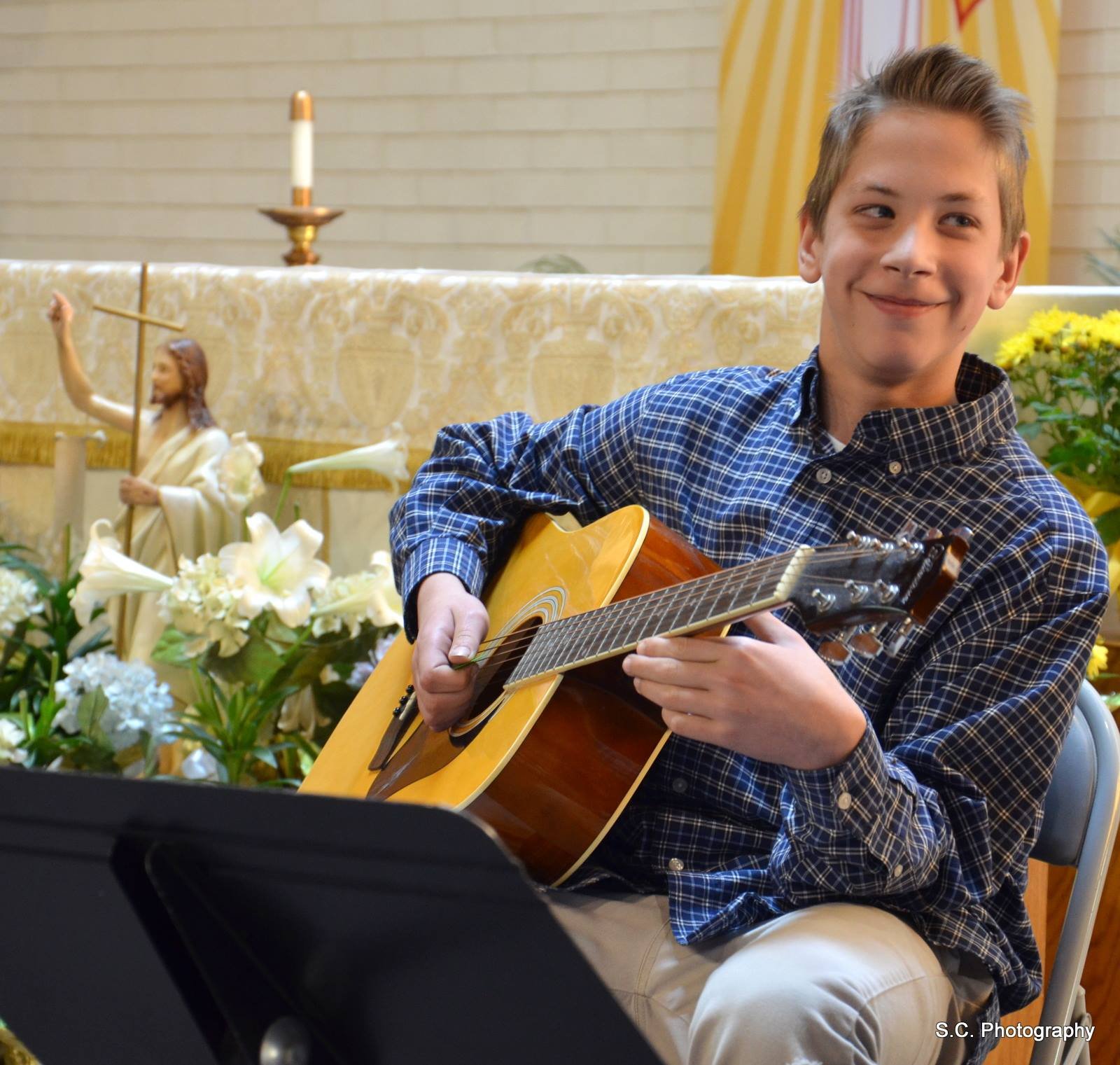 A smiling young boy playing an acoustic guitar in a church setting with religious decorations and flowers in the background.