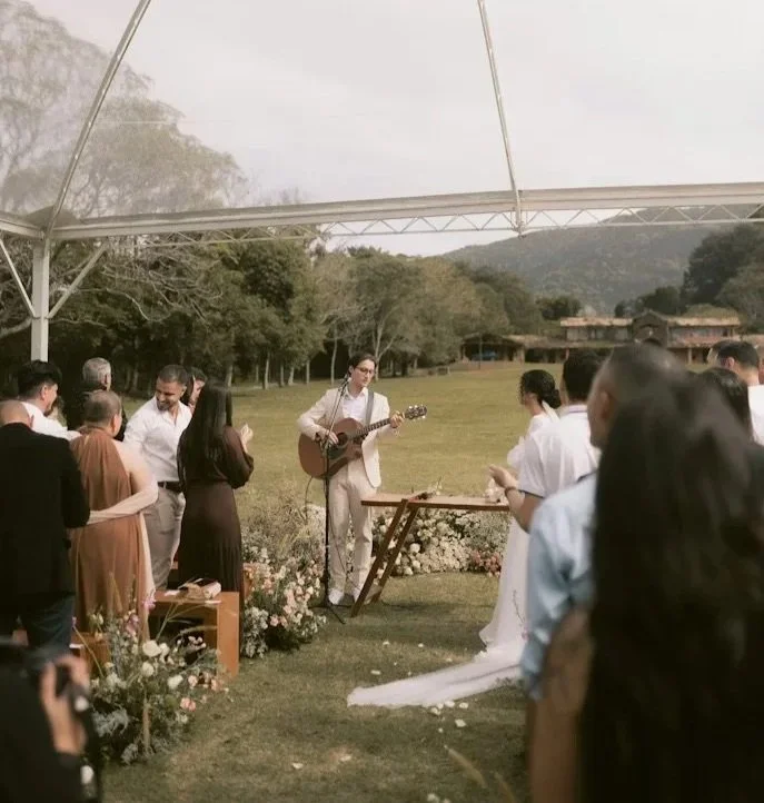 Man playing guitar and singing at an outdoor wedding ceremony, surrounded by guests, under a white canopy with a scenic background of trees and mountains.