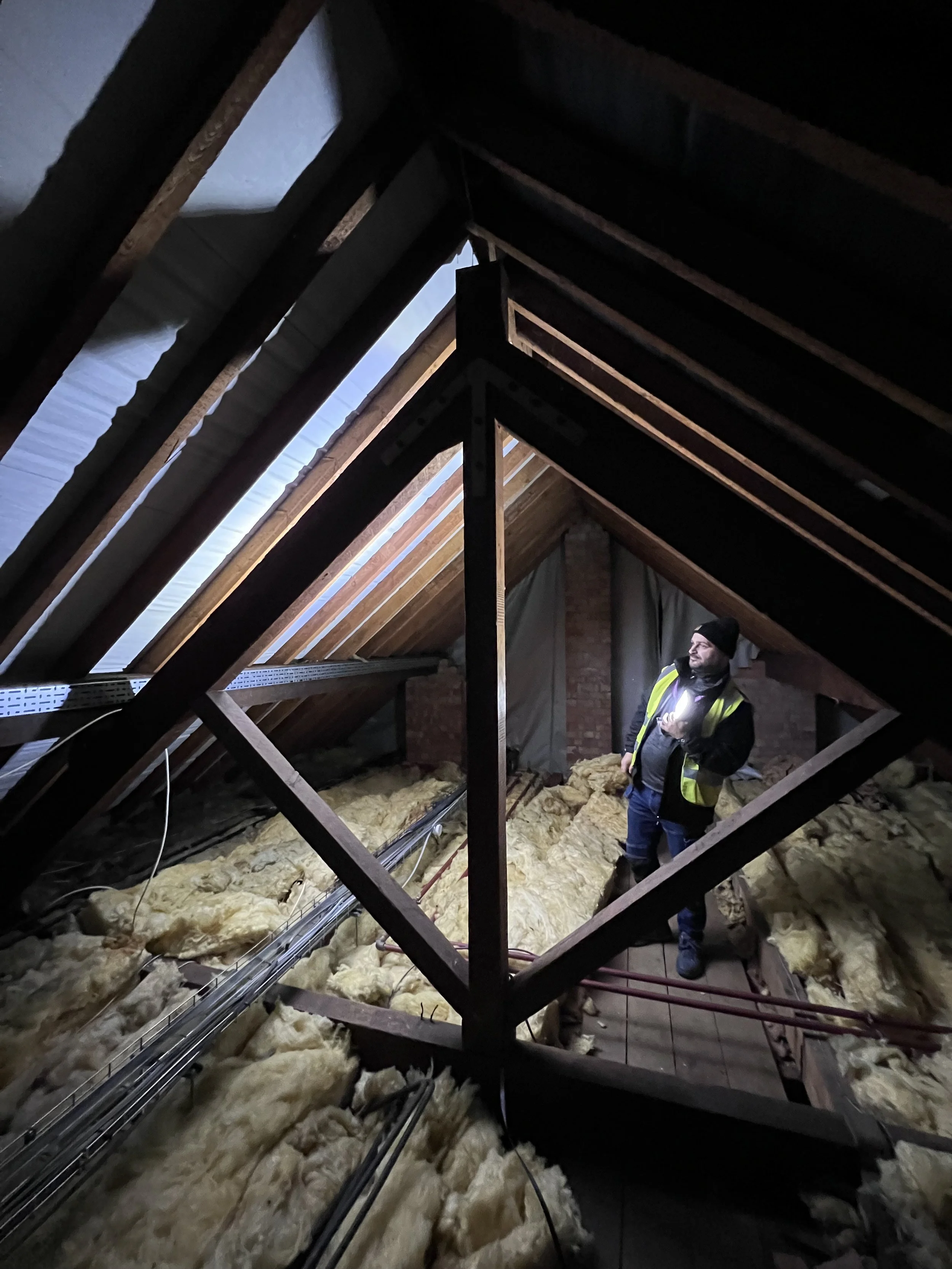 A man in a black beanie, black jacket, and yellow safety vest standing in an attic with exposed wooden beams, insulation, and electrical wiring.